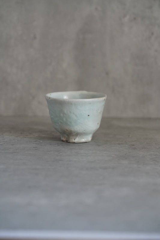 Small footed cup in pale green-tinged cream with fine speckles, on grey concrete.