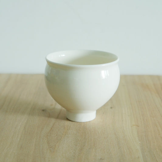 Plain white porcelain cup with a rounded body on a small foot, on a light wooden table.
