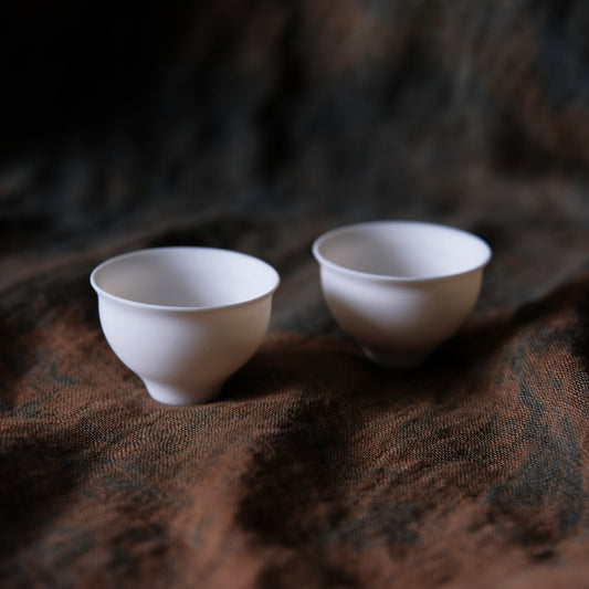 Two small white porcelain tea cups on low feet, on rumpled brown cloth.