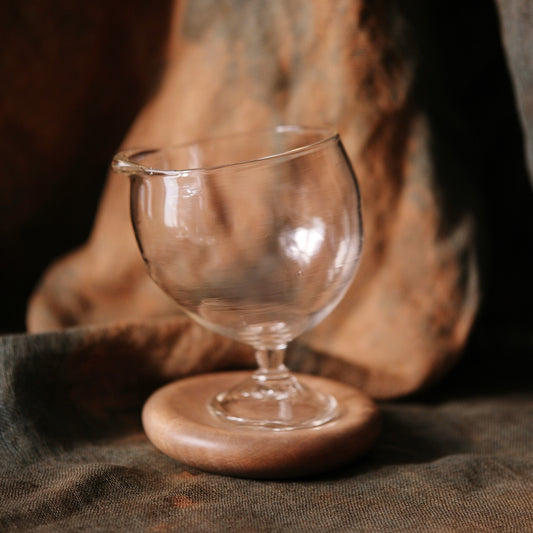 Clear glass goblet with a tilted bowl on a broad flat foot, against warm rust-coloured cloth.