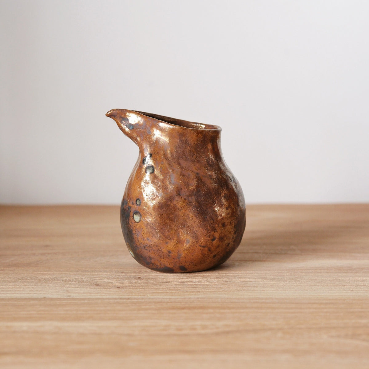 Small pouring vessel with a bronze metallic glaze and a sharply drawn spout, on a wooden table.
