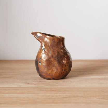 Small pouring vessel with a bronze metallic glaze and a sharply drawn spout, on a wooden table.