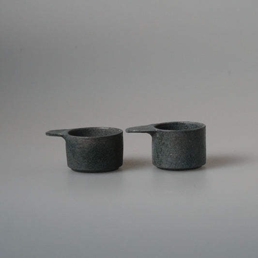 Two small dark ceramic scoops with short flat handles, side by side on pale grey.