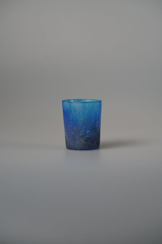 Small glass cup in deep blue speckled with green and white flecks, on pale grey.
