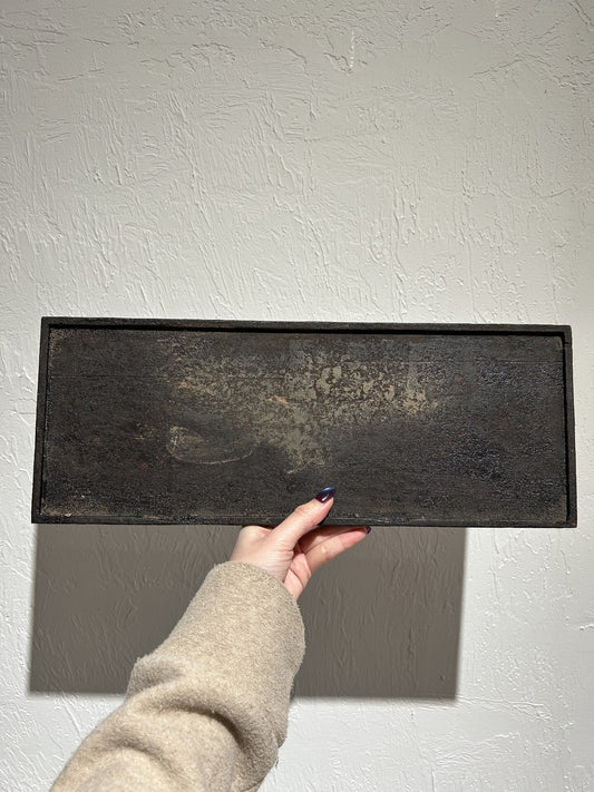 Large rectangular dish in black lacquer with a rusted, pitted surface, held against a white wall.
