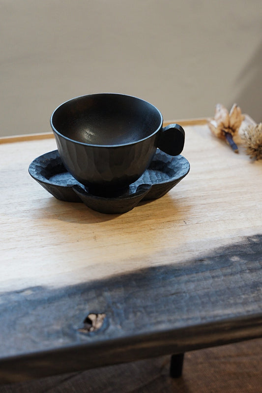 Small black lacquer cup with side ears, resting on a blue glazed saucer on a dark wooden table.