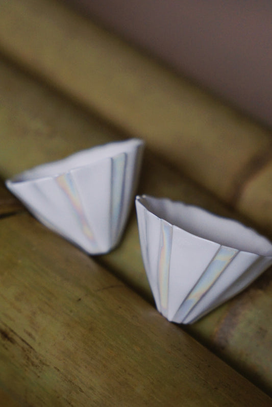 Two small angular white cups with pearly lustre stripes, resting on pale bamboo.