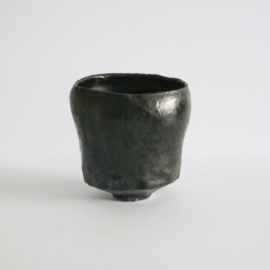 Black-glazed tea bowl on a small foot, its rim dipping in a soft wave, lit against white.