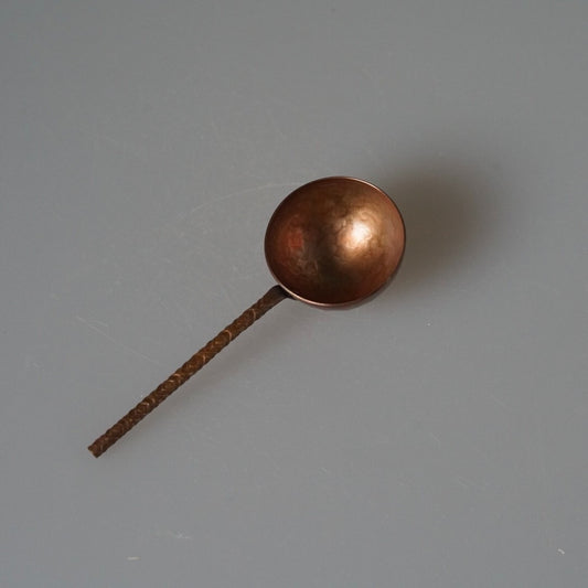 Copper coffee scoop with a deep round bowl and a slim hammered handle, on pale grey.