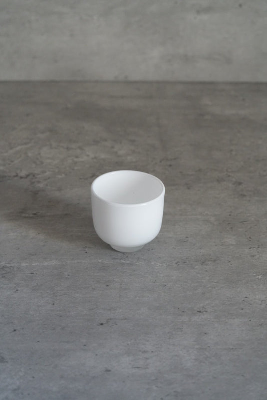 Small opaque milk-white glass cup with straight sides, on a pale grey floor.