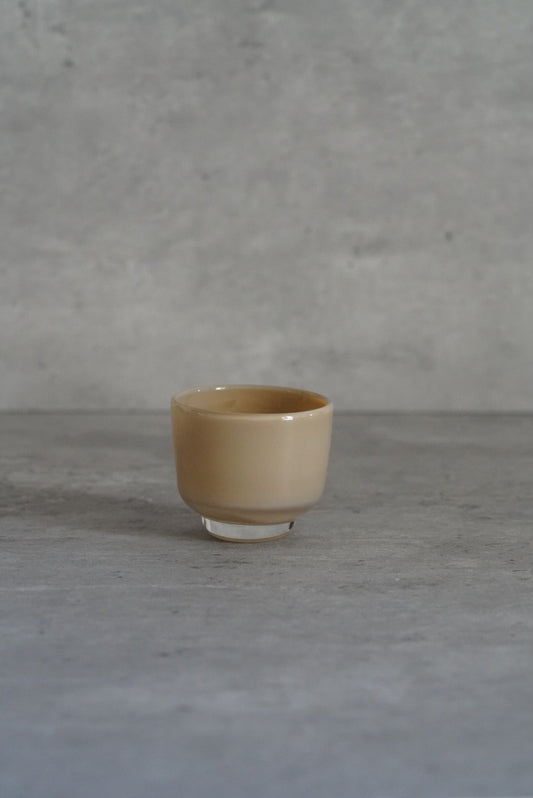 Small opaque glass cup in soft sand yellow on a low foot, on a pale grey floor.