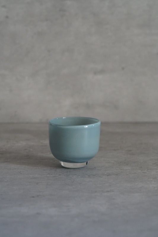 Small opaque glass cup in soft duck-egg blue on a low foot, on a pale grey floor.