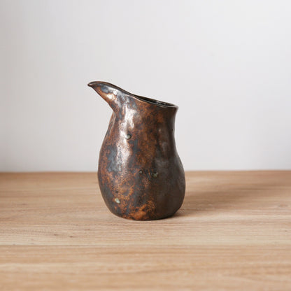 Bronze-glazed pouring jug with a full belly and a long drawn lip, on a wooden table.