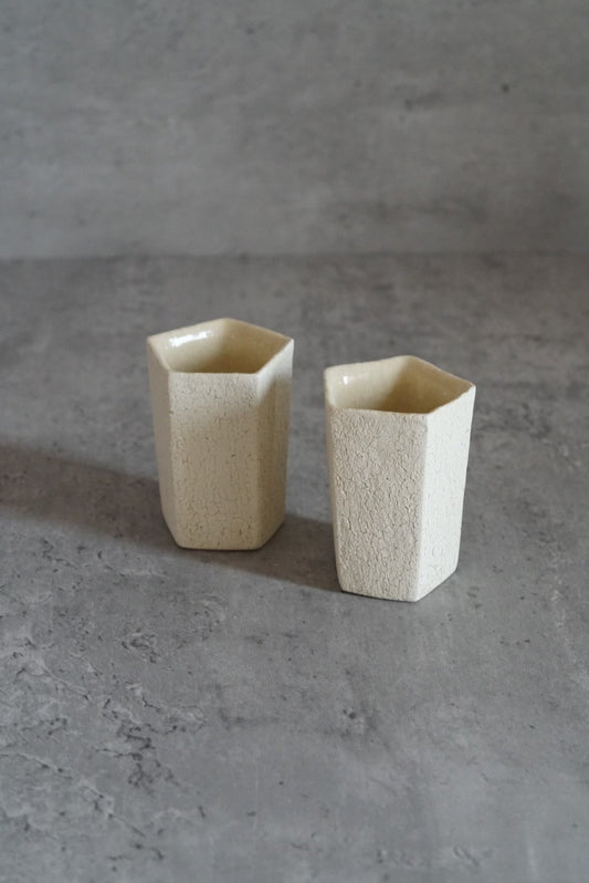 Two small faceted cups in cream and white with cut flat sides, on grey concrete.