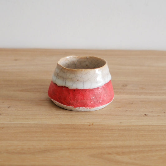 Small tapered cup banded in bright red below a rough white rim, on a light wooden table.