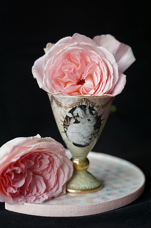Tall painted cup on a gold pedestal foot, standing on a pale dotted saucer beside two pink garden roses.