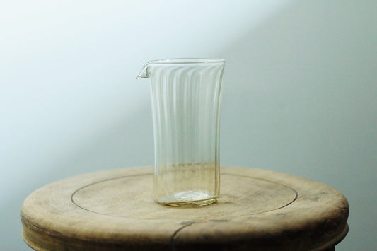 Tall ribbed glass pourer with a drawn lip and a faint amber tint, on a pale wooden stump.