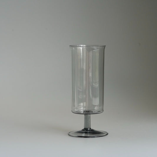 Tall clear glass on a slim smoky grey stem and round foot, on a pale ground.