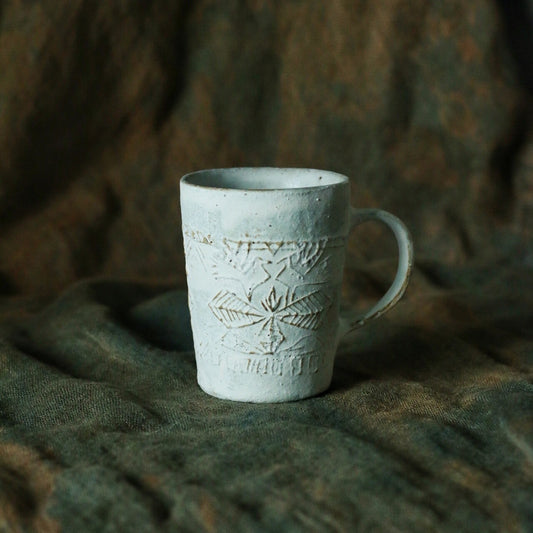 Pale green mug incised with faint flowers and lettering, with a slim handle, on green cloth.