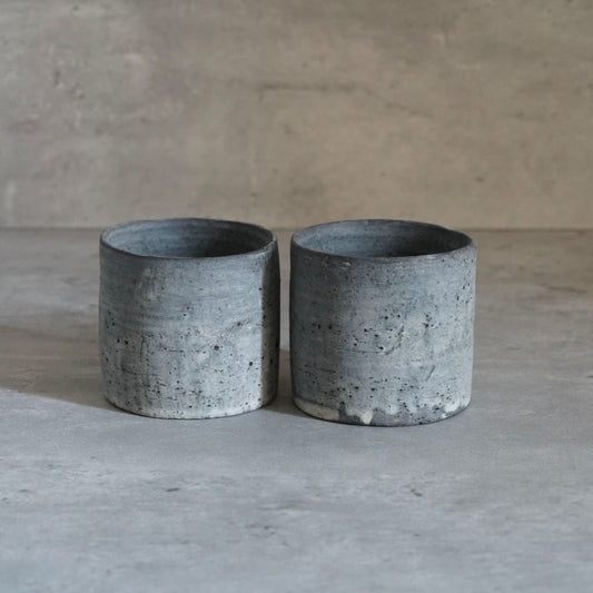 Two straight-sided cups in mottled blue-grey with rough pitted surfaces, side by side.