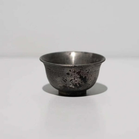 Small footed cup in dark tarnished silver-grey with a mottled, weathered surface.