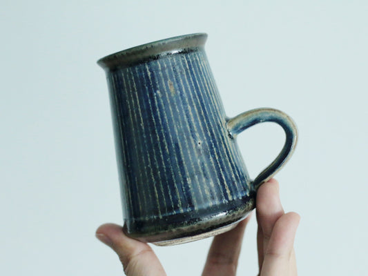 Tall deep blue mug with vertical ribbing and a small loop handle, held up in a hand.