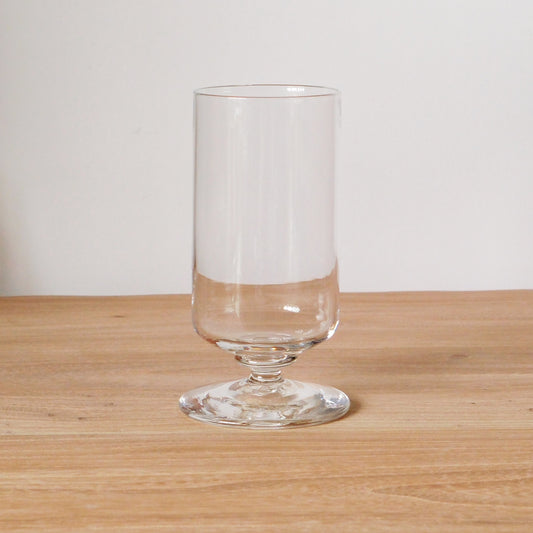 Tall clear glass on a stepped foot, its straight bowl catching a soft band of light.