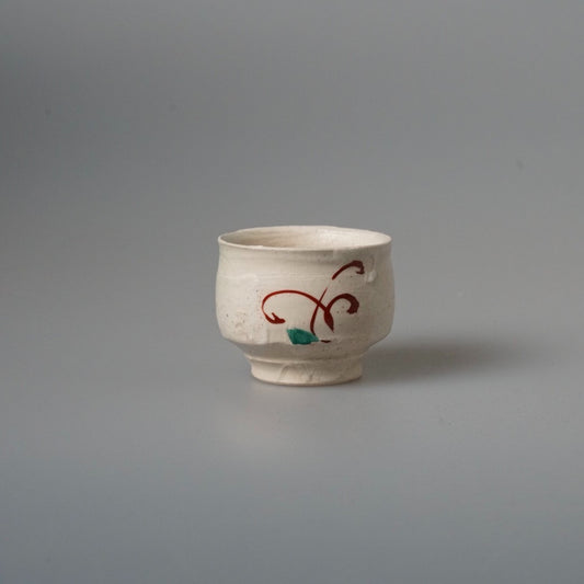 Small cream cup painted with a loose red scrolling line and a green leaf mark.