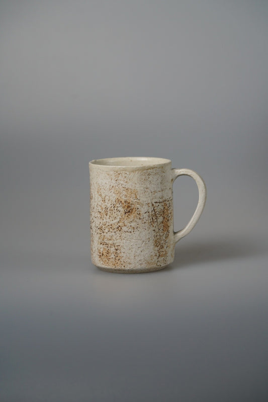 Tall cream mug marked with rust blushes and a slim loop handle, on pale grey.