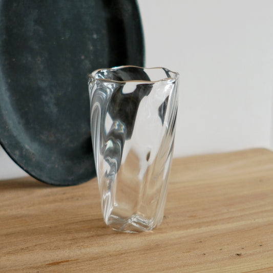 Tall clear glass with a twisted, softly folded body catching bright reflections, on wood.