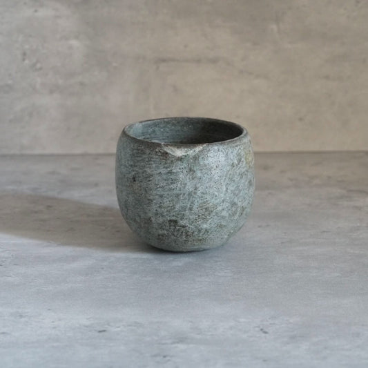 Round cup in mottled grey-green with a coarse pitted surface, on grey concrete.