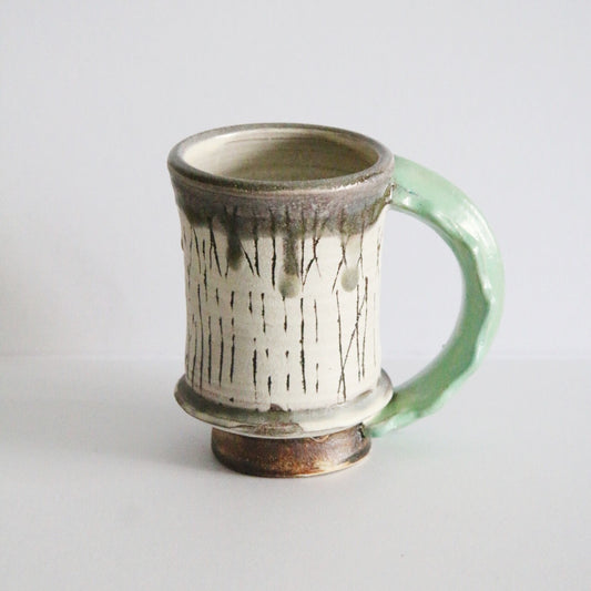 Tall cream mug streaked with dark brush lines, with a pale green handle and a bronze rim.
