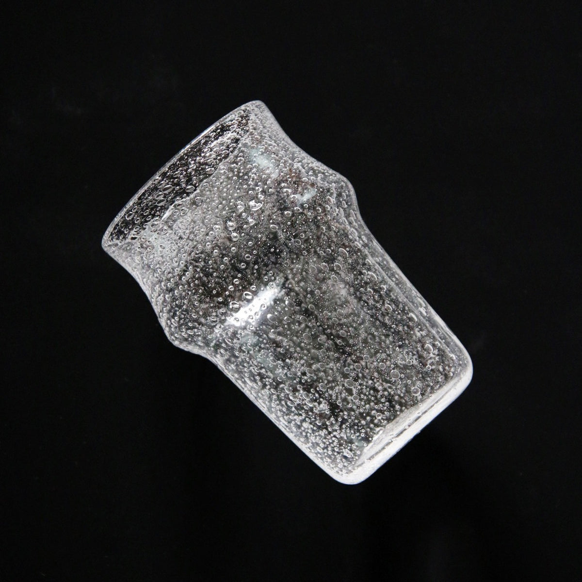 Tall waisted clear glass tumbler dense with tiny bubbles, tilted against a black ground.