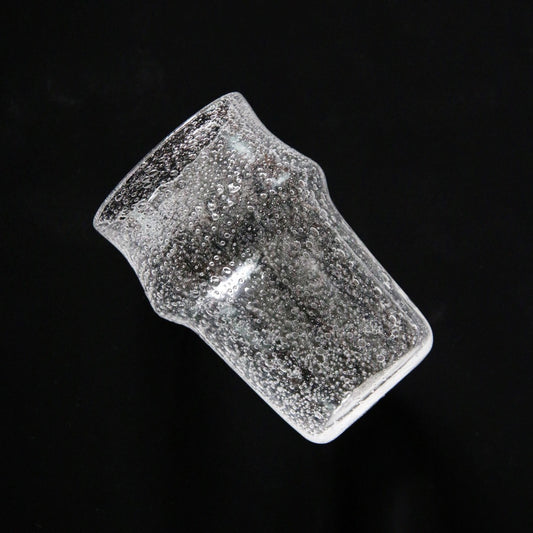 Tall waisted clear glass tumbler dense with tiny bubbles, tilted against a black ground.