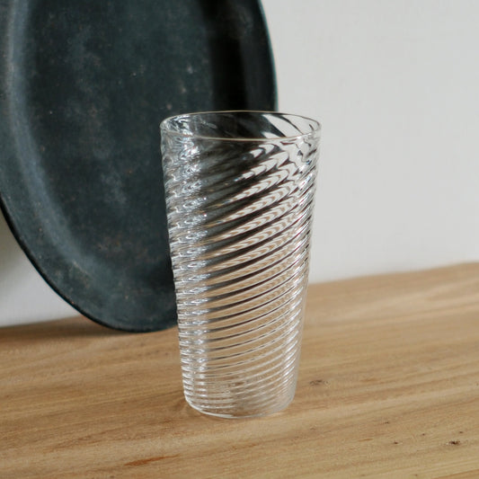 Tall clear glass tumbler with fine spiralling ribs running up its sides, on a wooden table.