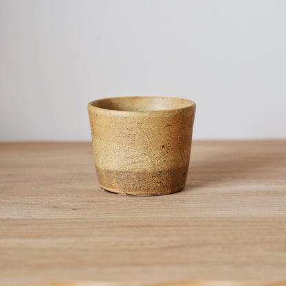 Straight-sided cup in warm sandy brown with a rough grainy surface, on a wooden table.