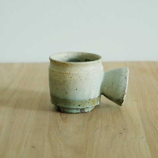 Speckled cream cup with a soft green glaze low on its body, beside a second cup lying on its side.