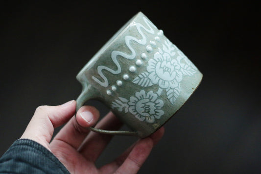 Pale green mug held in a hand, carved and painted with white flowers and a beaded rim.