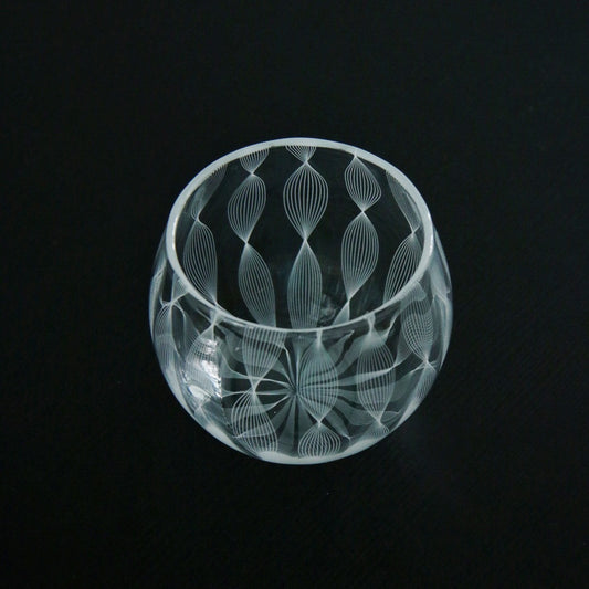 Rounded clear glass cup cut with a fine lace pattern of teardrops and rays, lit against black.