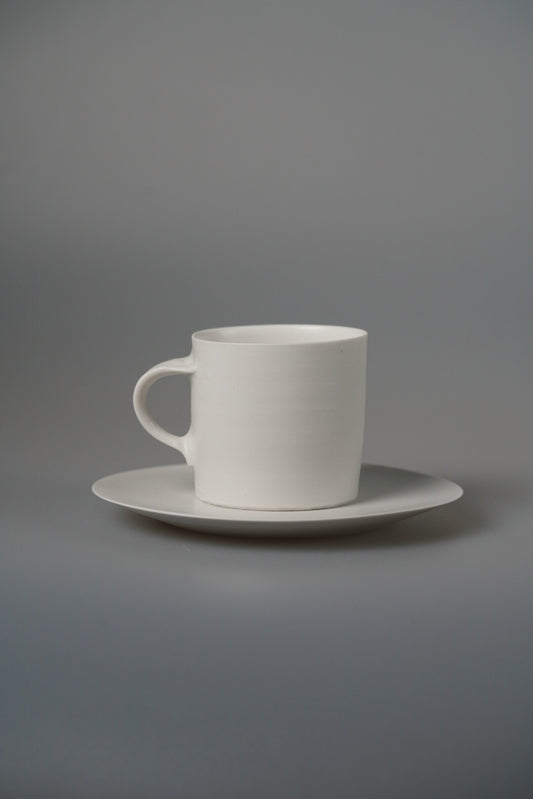 Plain white porcelain mug with a squared handle, standing on a matching white saucer.