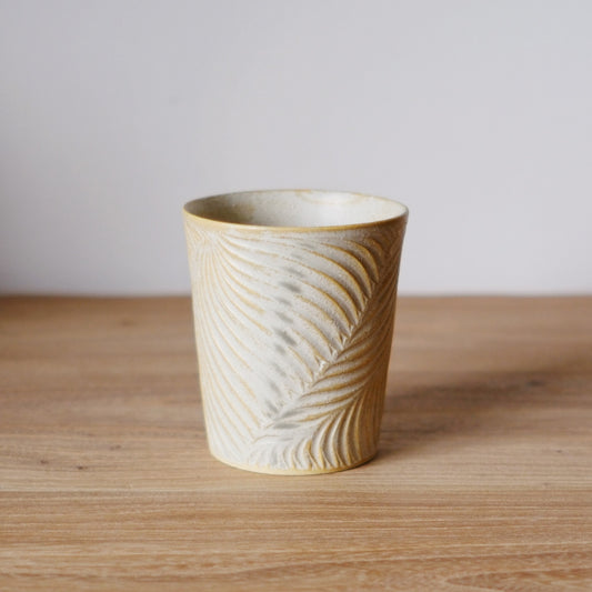 Tapered cream cup carved all over with fine feathered lines, on a light wooden table.