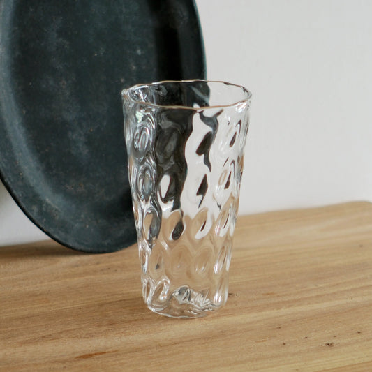 Tall clear glass tumbler with a rippled dimpled surface, on a light wooden table.