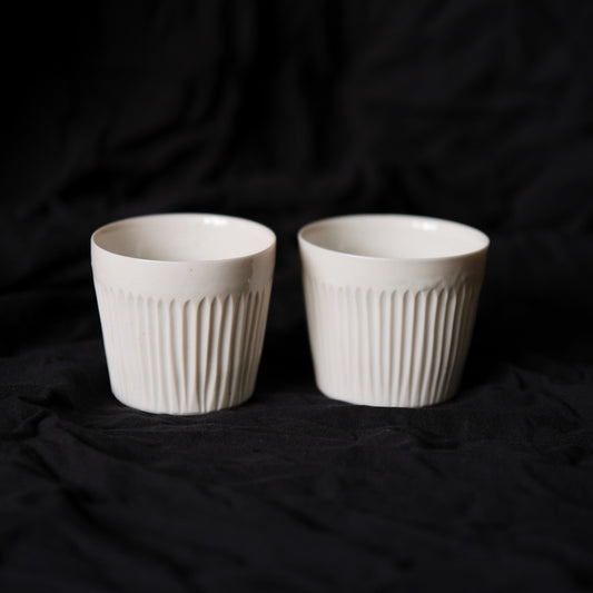 Two white porcelain cups carved with close vertical lines, side by side on black cloth.
