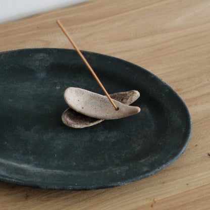 Two small pebble-shaped incense stands, one pale and one dark, holding a stick on a black plate.
