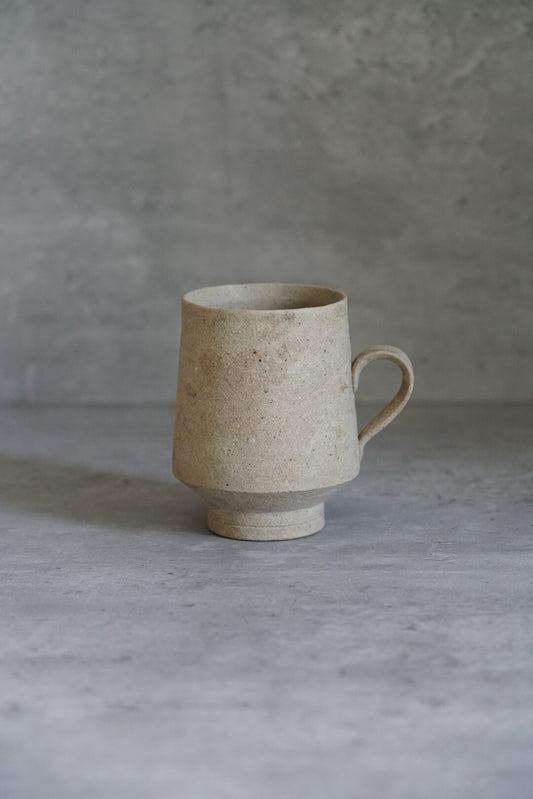 Tall footed cream mug with a speckled matte surface and a small loop handle.