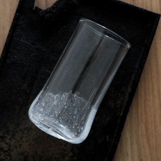 Tall clear glass tumbler filled with fine trapped bubbles, lying on its side on a dark board.