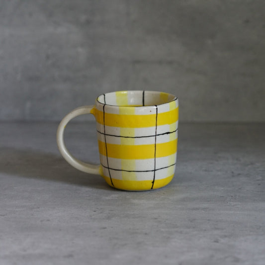 White mug painted with a bold yellow check grid and a thin white loop handle.