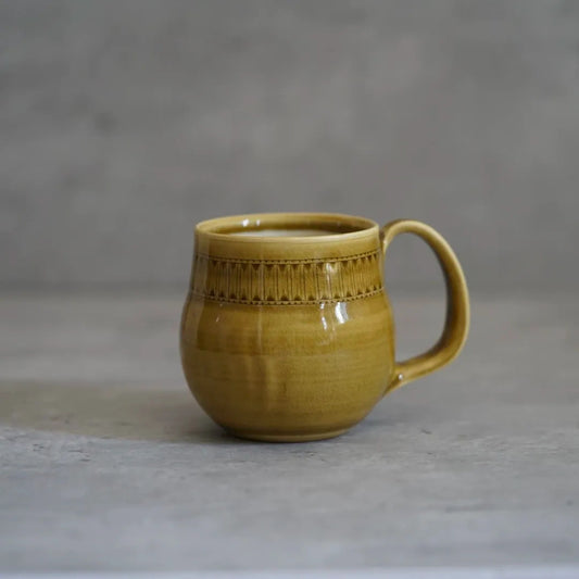 Round mustard yellow mug with a band of stamped marks below the rim and a loop handle.