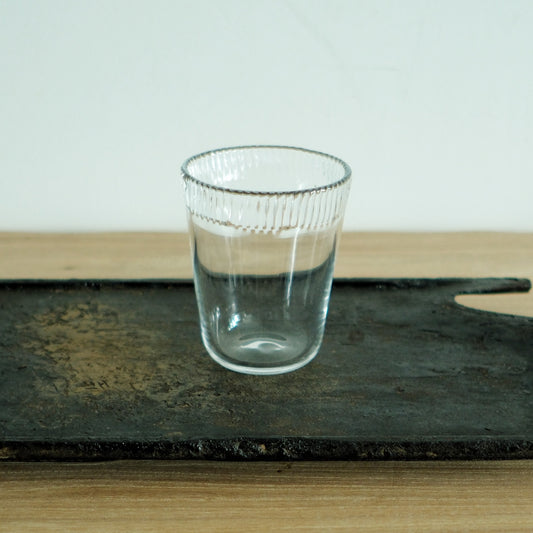 Tall clear glass tumbler with fine ribbing near the rim, on a dark weathered board.