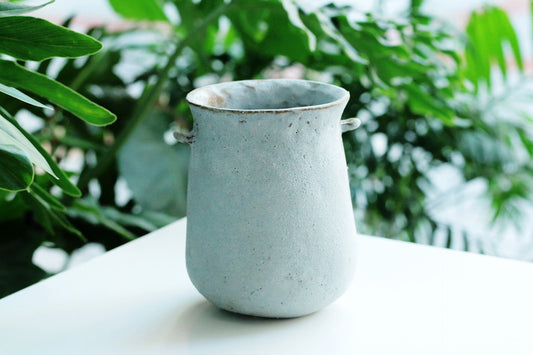 Small two-handled vase in speckled sage grey, standing on white beside green foliage.